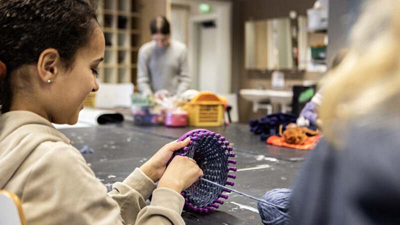 A child smiles while knitting a purple-blue basket in a craft class. In the background, another child is focused on their own project.