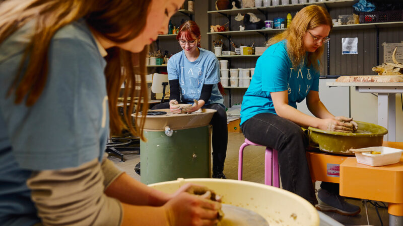 Three people sitting at pottery wheels, focused on their work. They are wearing blue shirts and working in a pottery studio.