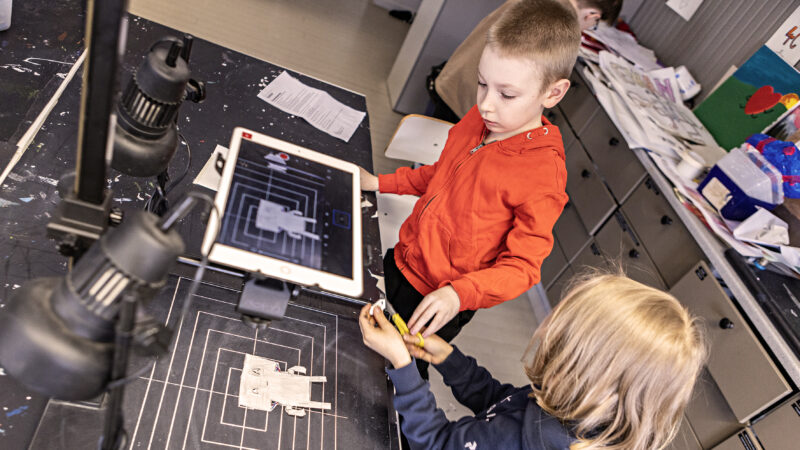 Two children working together in a classroom. They are using a tablet and looking at drawings on a black table.