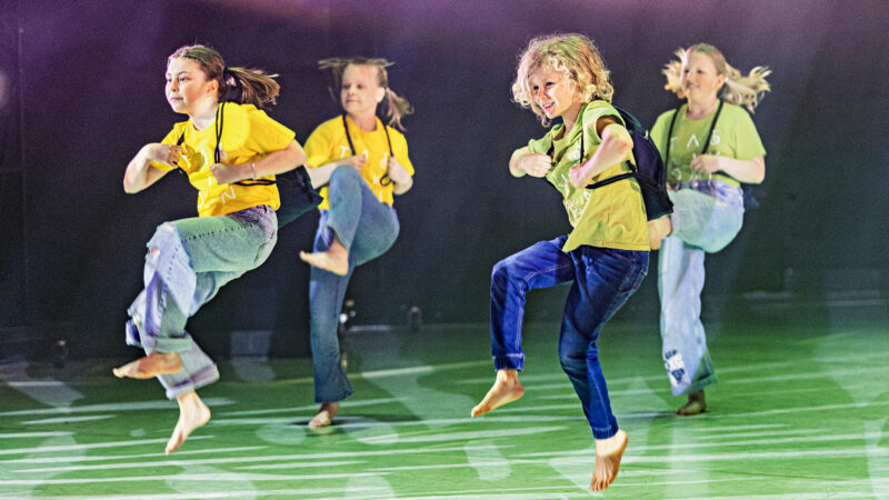 Four children energetically performing on a dance stage, wearing yellow shirts and jeans.