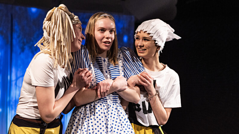 Three young actors perform on stage. The person in the middle is wearing a blue and white polka dot dress, and the two other individuals are wearing t-shirts and holding her hands.