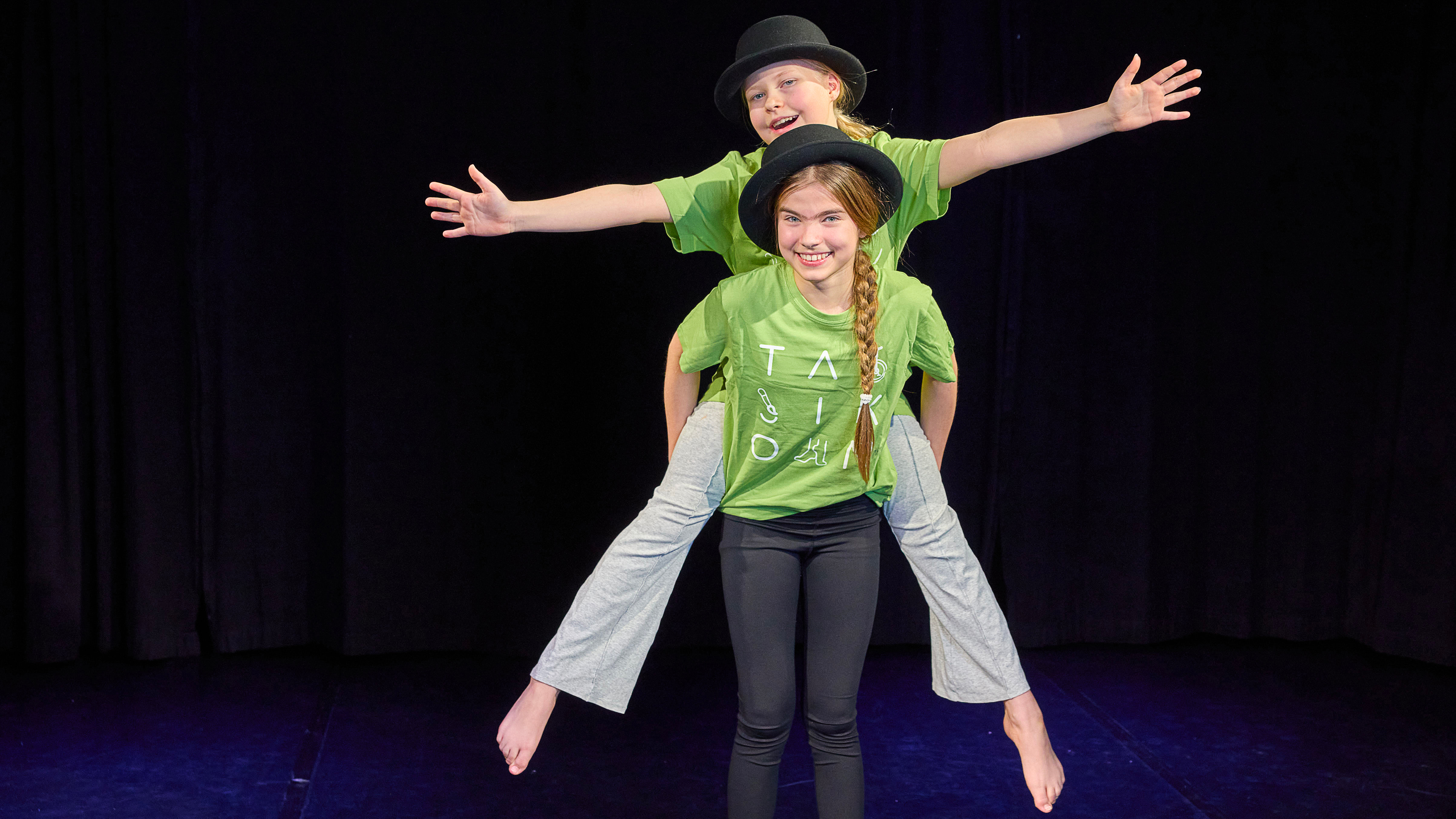 Two children performing on stage. One child is standing on the shoulders of the other, both wearing green shirts and black hats.