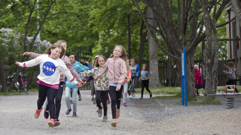 Children running happily on a playground with green trees in the background.