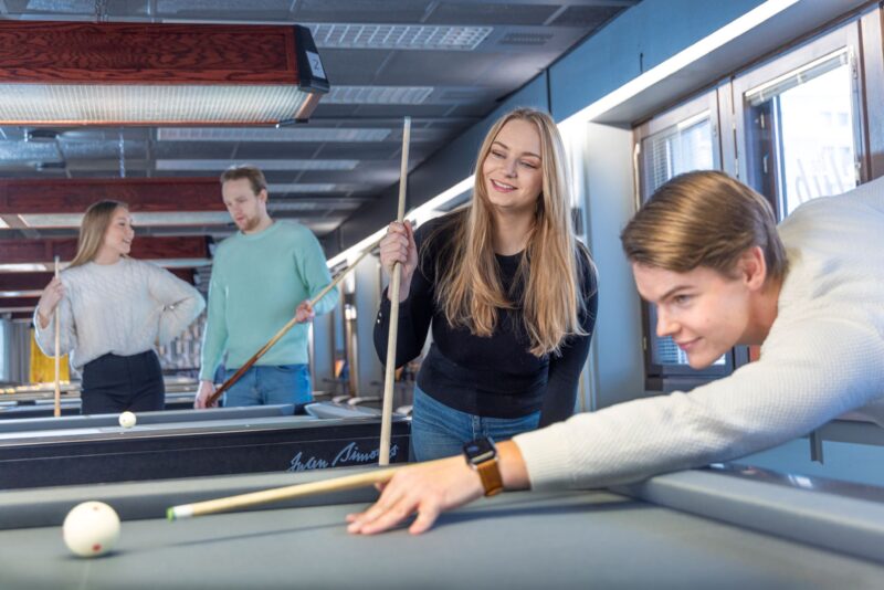 Tre personer spelar biljard inomhus. I förgrunden siktar en person med en kö, i mitten ler en person, och i bakgrunden står två personer och pratar.