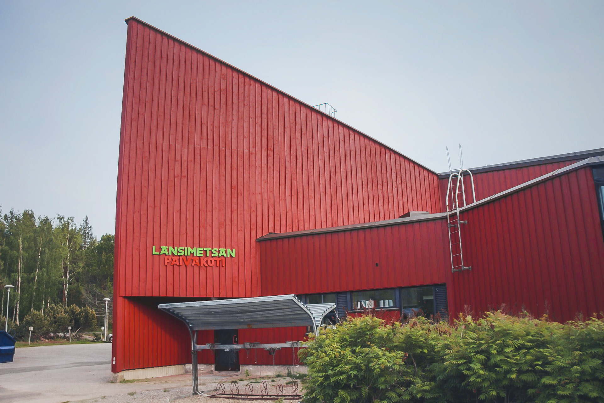 Modern röd byggnad med text 'Länsimetsän päiväkoti' och gröna träd runt omkring.