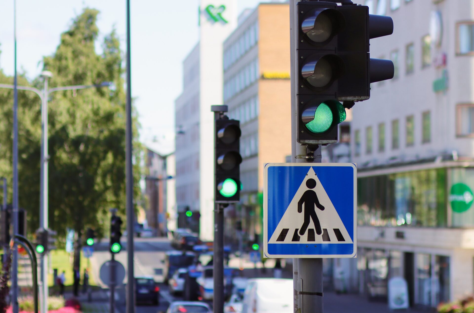 Kuva vilkkaasta kadusta, jossa on liikennevalot ja jalankulkijan varoitusmerkki. Liikennevalot näyttävät vihreää.