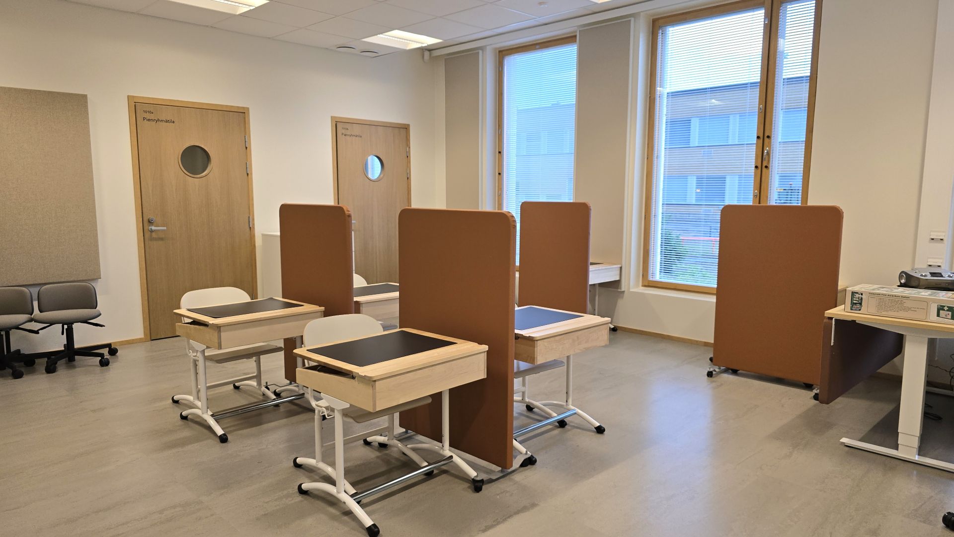 Klassrum med fyra skrivbord med avdelare och stora fönster på höger sida.