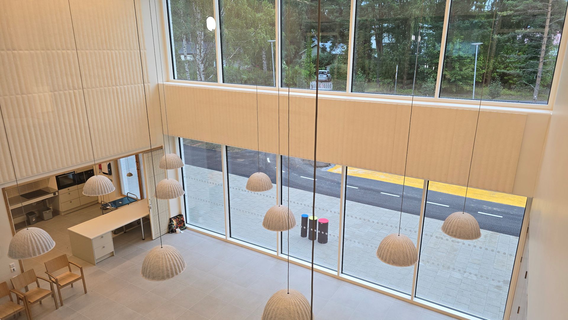 En modern lobby med stora fönster, hängande lampor och några stolar. Träd och en väg syns utanför.