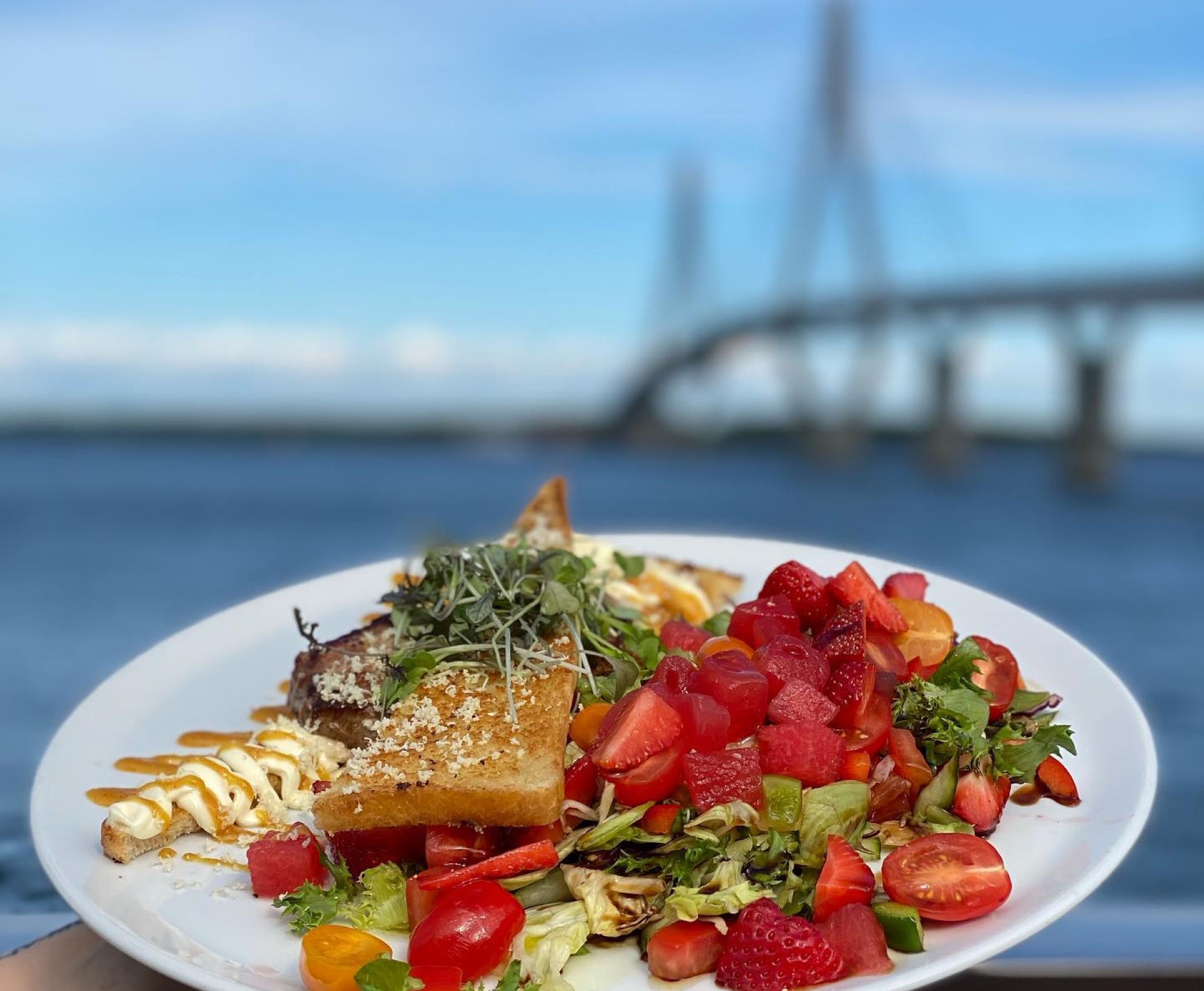 En tallrik med färgglad mat innehållande sallad, vattenmelon, jordgubbar, rostat bröd och dressing, med en vacker bro och blå himmel i bakgrunden.