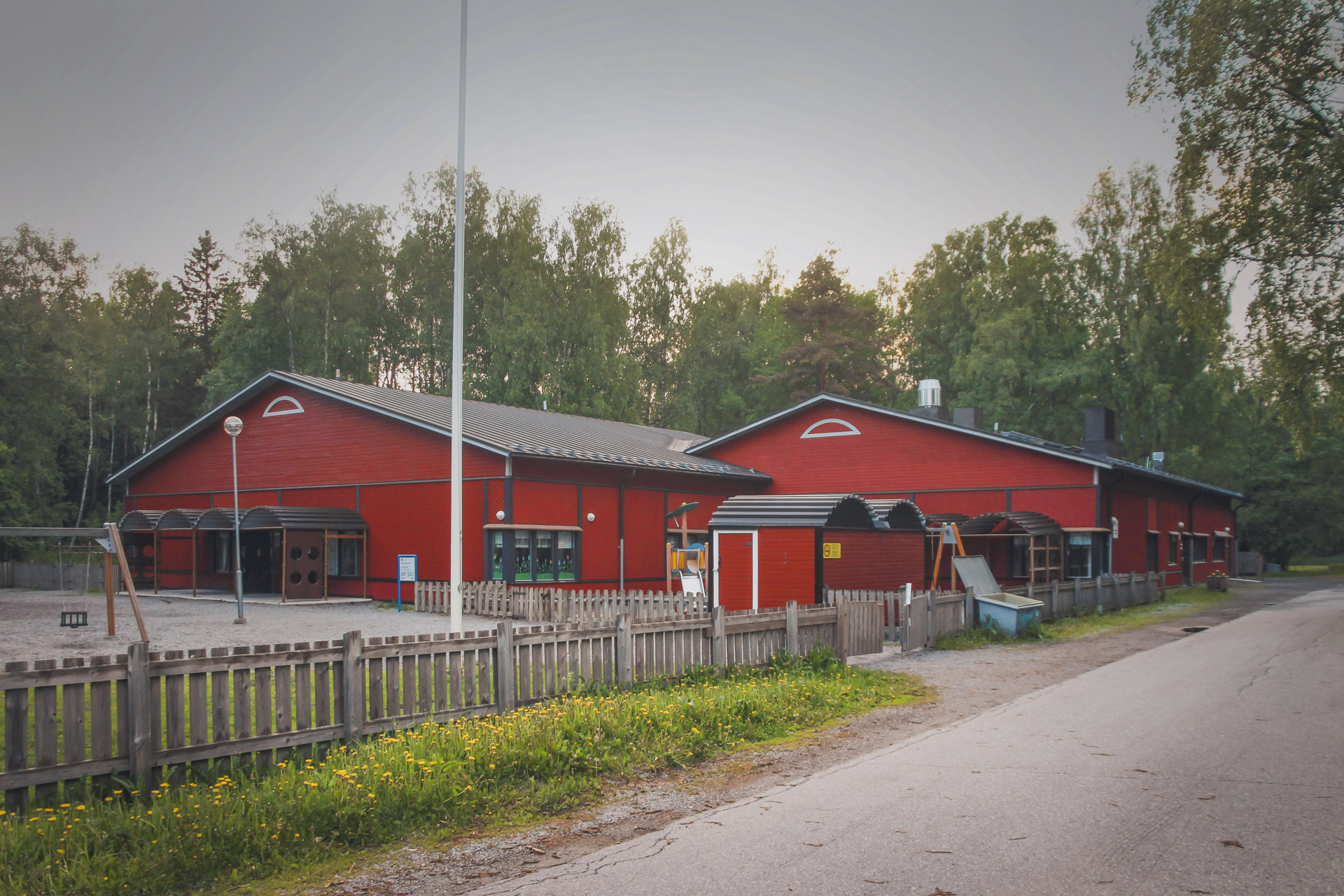 Röd byggnad omgiven av träd, gård och trästaket i förgrunden.