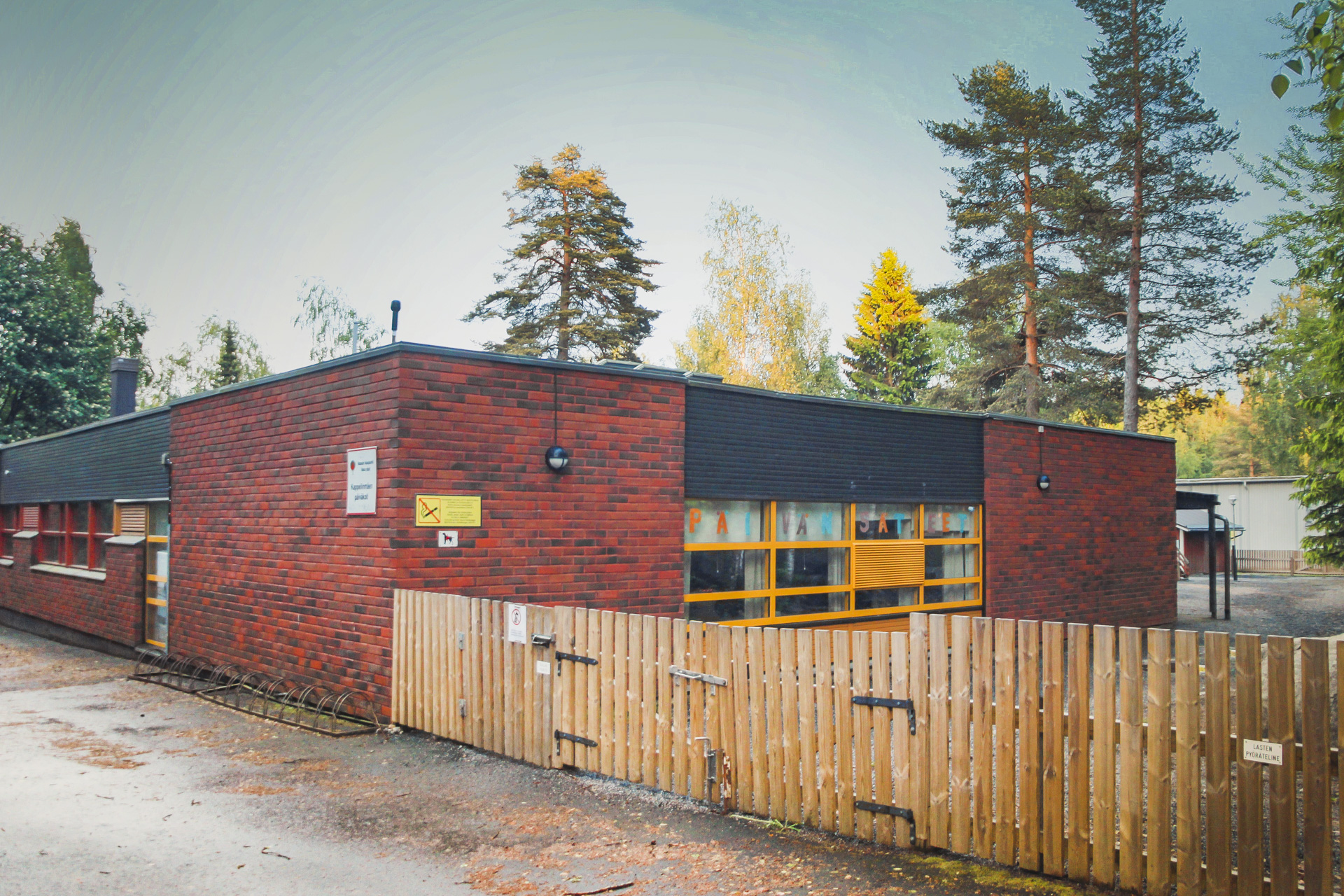 Tegelbyggnad med stora fönster och trästaket i förgrunden. Träd och små byggnader i bakgrunden.