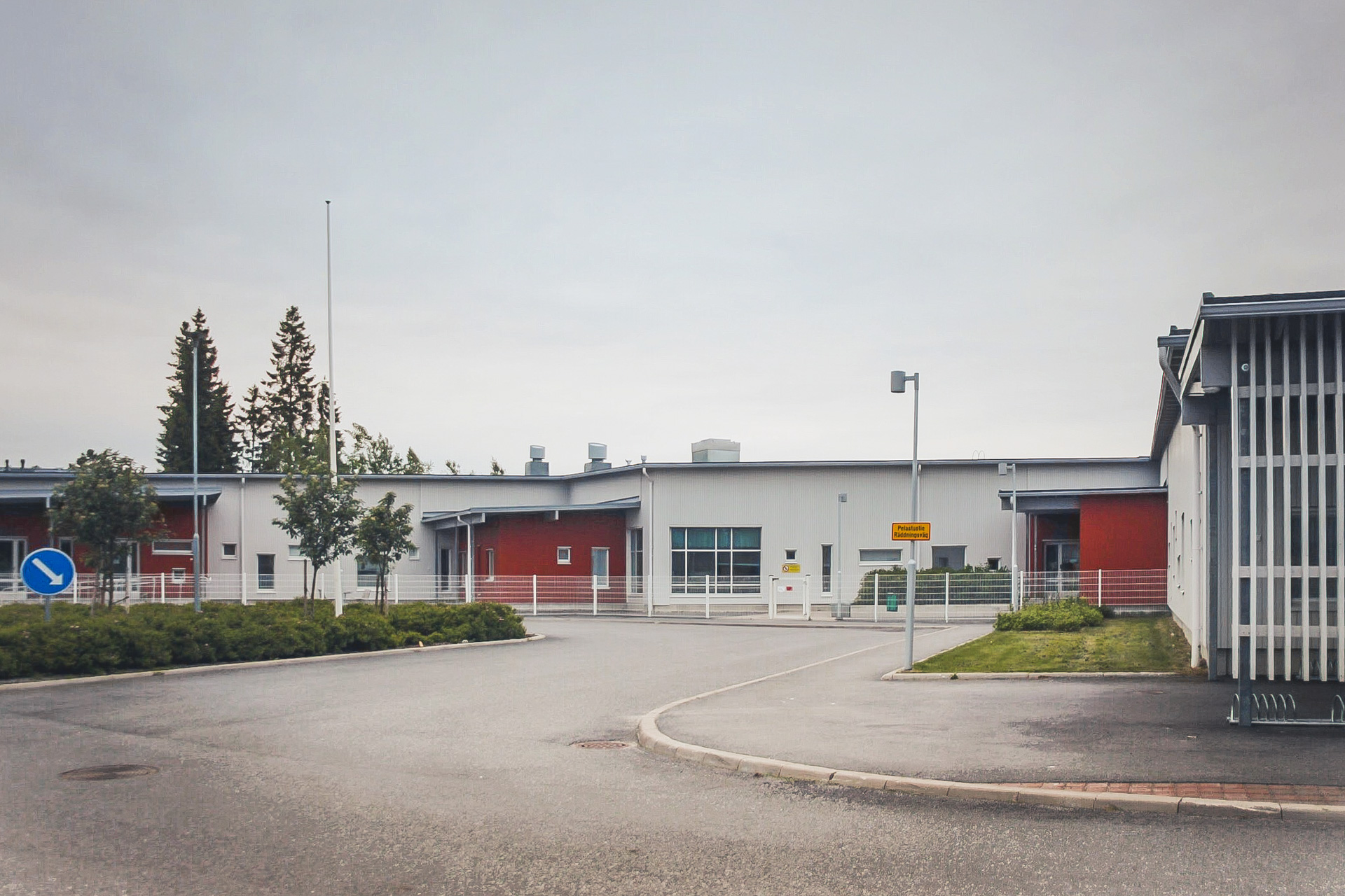 Daghemsbyggnad med röda och grå väggar omgiven av stor asfaltplan och gräsmatta. Träd i bakgrunden.
