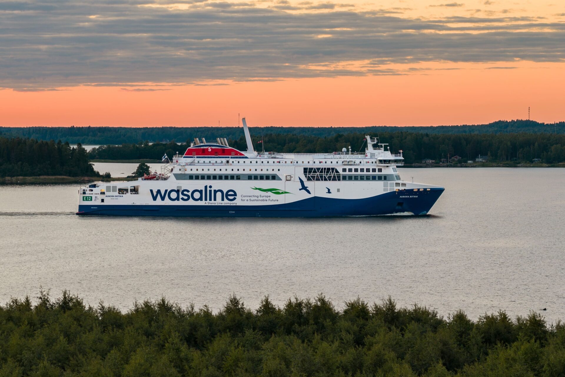 Wasalinen laiva kulkee merellä auringonlaskun aikaan. Taustalla näkyy metsäinen ranta.