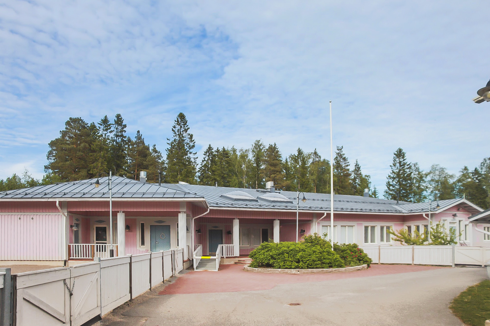 Ljusrosa enplansbyggnad omgiven av skog. Blå himmel i bakgrunden.