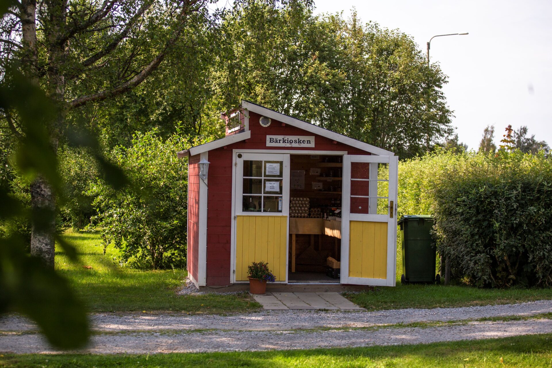 En röd träkiosk med gula dörrar på en solig landsbygd, med en krukväxt framför.