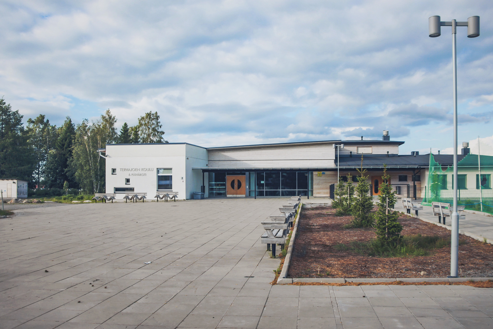 Modern skolbyggnad med stor gård och bänkar, omgivet av skog.