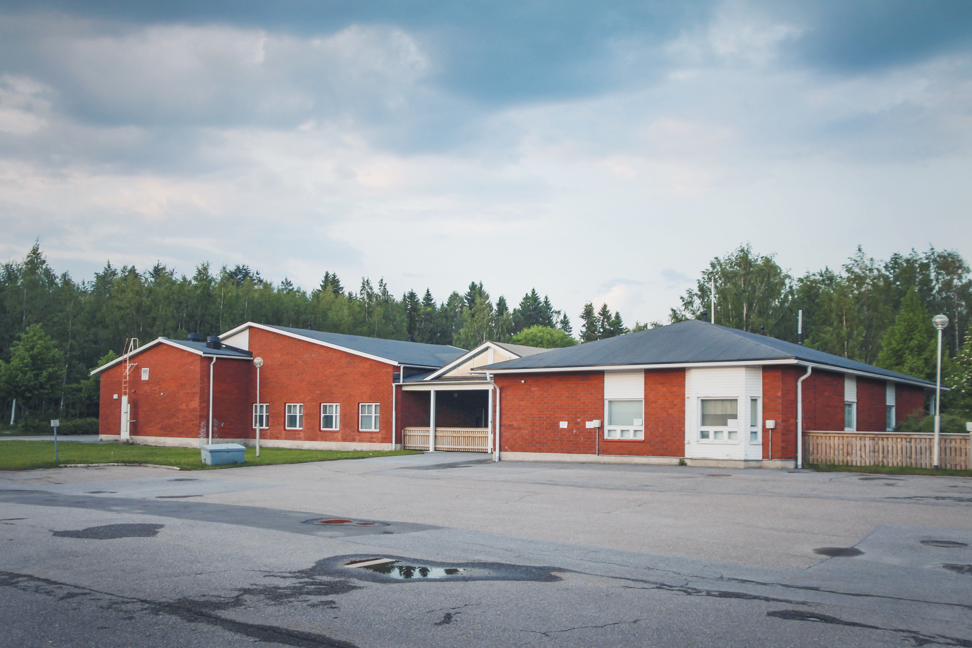 Röd tegelbyggnad med grått tak på en tom asfalterad gård. Skog i bakgrunden och molnig himmel.