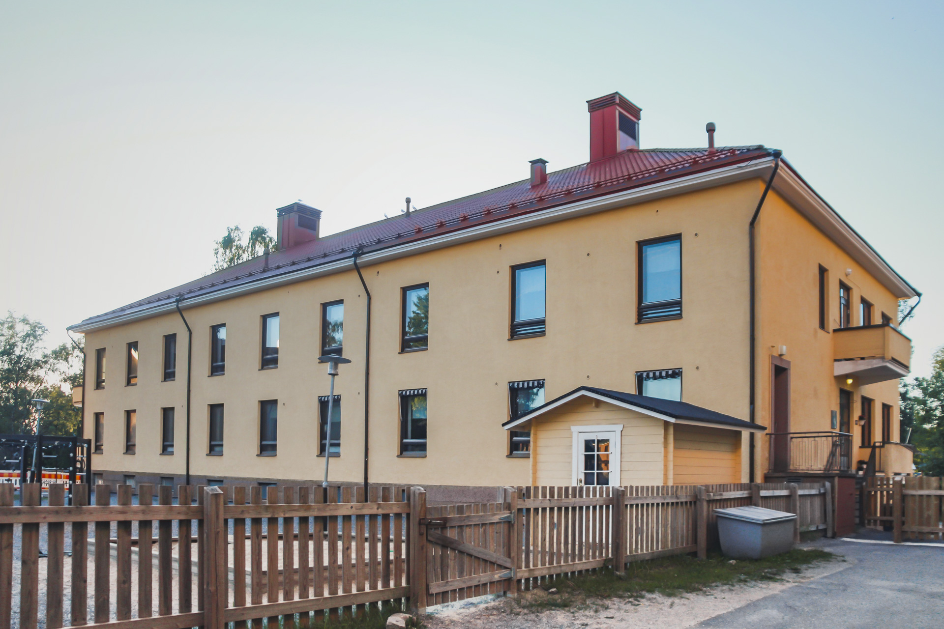 Gul tvåvåningsbyggnad med rött tak och trästaket framför.