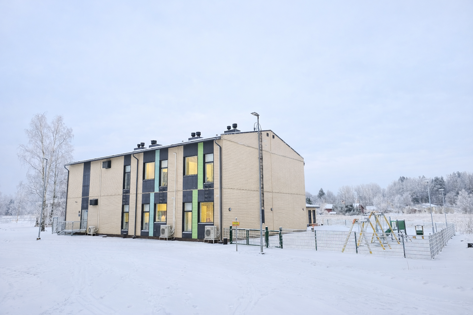 Tvåvåningsbyggnad i vinterlandskap med lekplats och snöklädda träd.