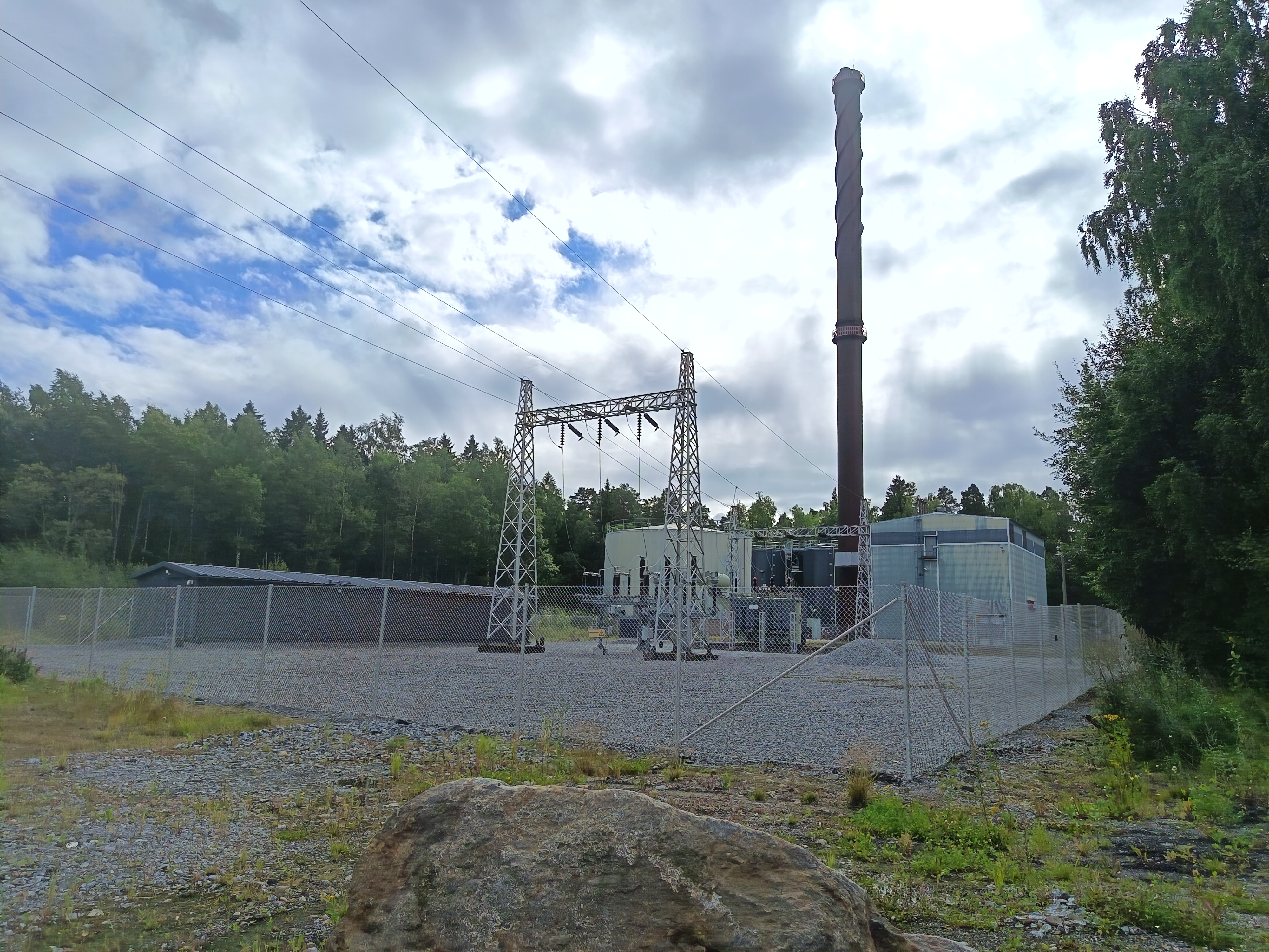 Kuva voimalaitoksesta, jossa on metallinen savupiippu ja sähkönjakelurakenteita aidatulla alueella. Taustalla näkyy metsäinen maisema ja pilvinen taivas.