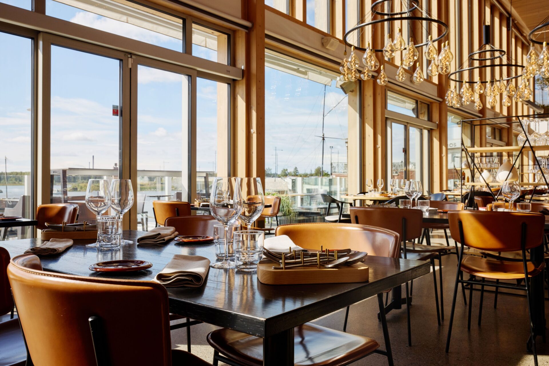 En restaurang med stora fönster, träinredning och ljuskronor. Borden är dukade med vinglas och servetter.