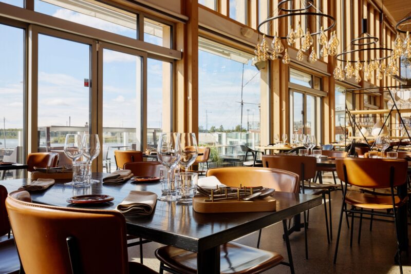 Ravintola, jossa on suuret ikkunat, puiset sisustuselementit ja kattokruunut. Pöydillä on viinilaseja ja lautasliinoja.