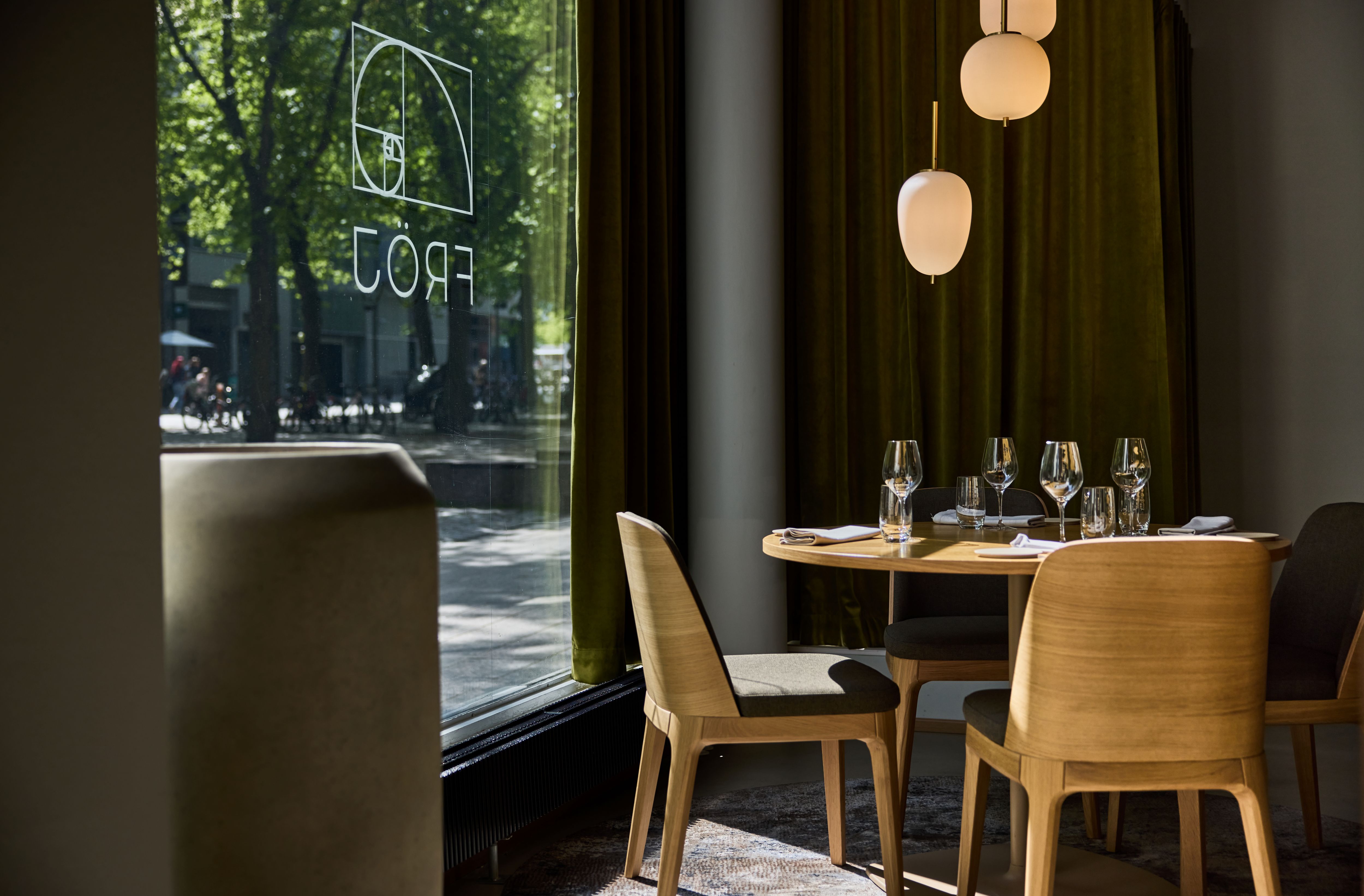 Restauranginteriör med ett runt träbord och fyra stolar. Bordet är dukat med vinglas och tallrikar, och en grön gatubild syns genom fönstret. Inredningen har varma färger och hängande lampor.