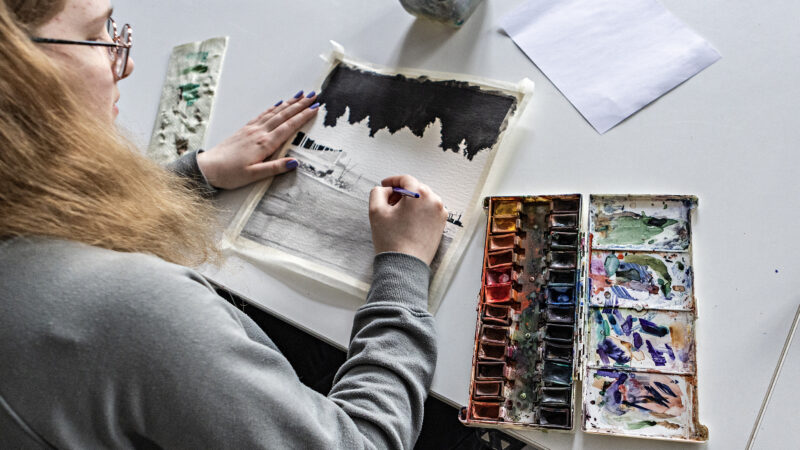 A person painting a landscape with trees using watercolors on paper.