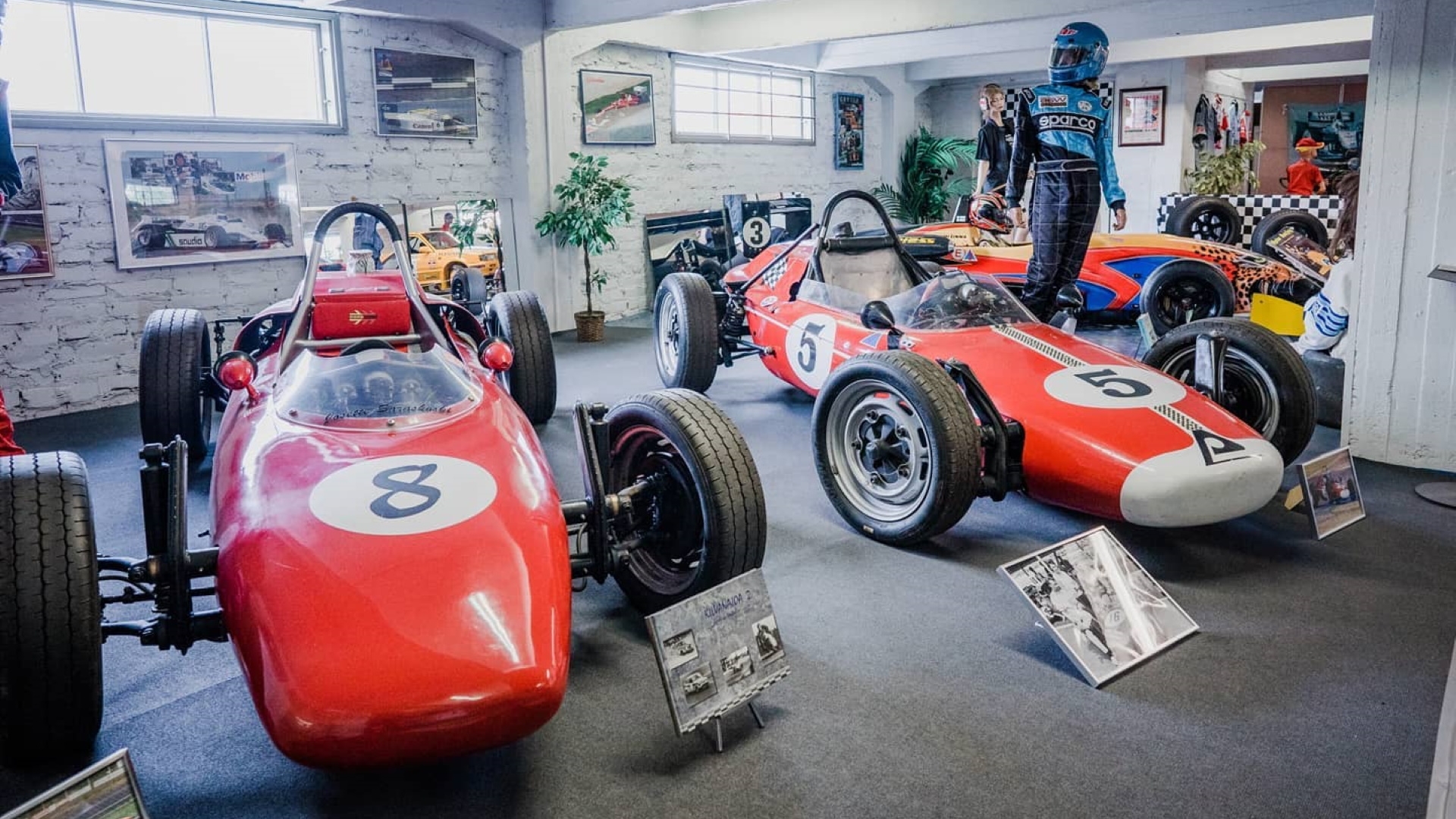 Vintage-kilpa-autot museossa, kaksi punaista autoa numerolla 8 ja 5, sekä mannekiini kilpa-ajajan asussa.
