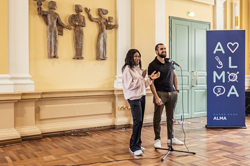 Two people stand at a microphone in a room with yellow walls and decorative sculptures. Next to them is a blue sign with the text 'ALMA'.