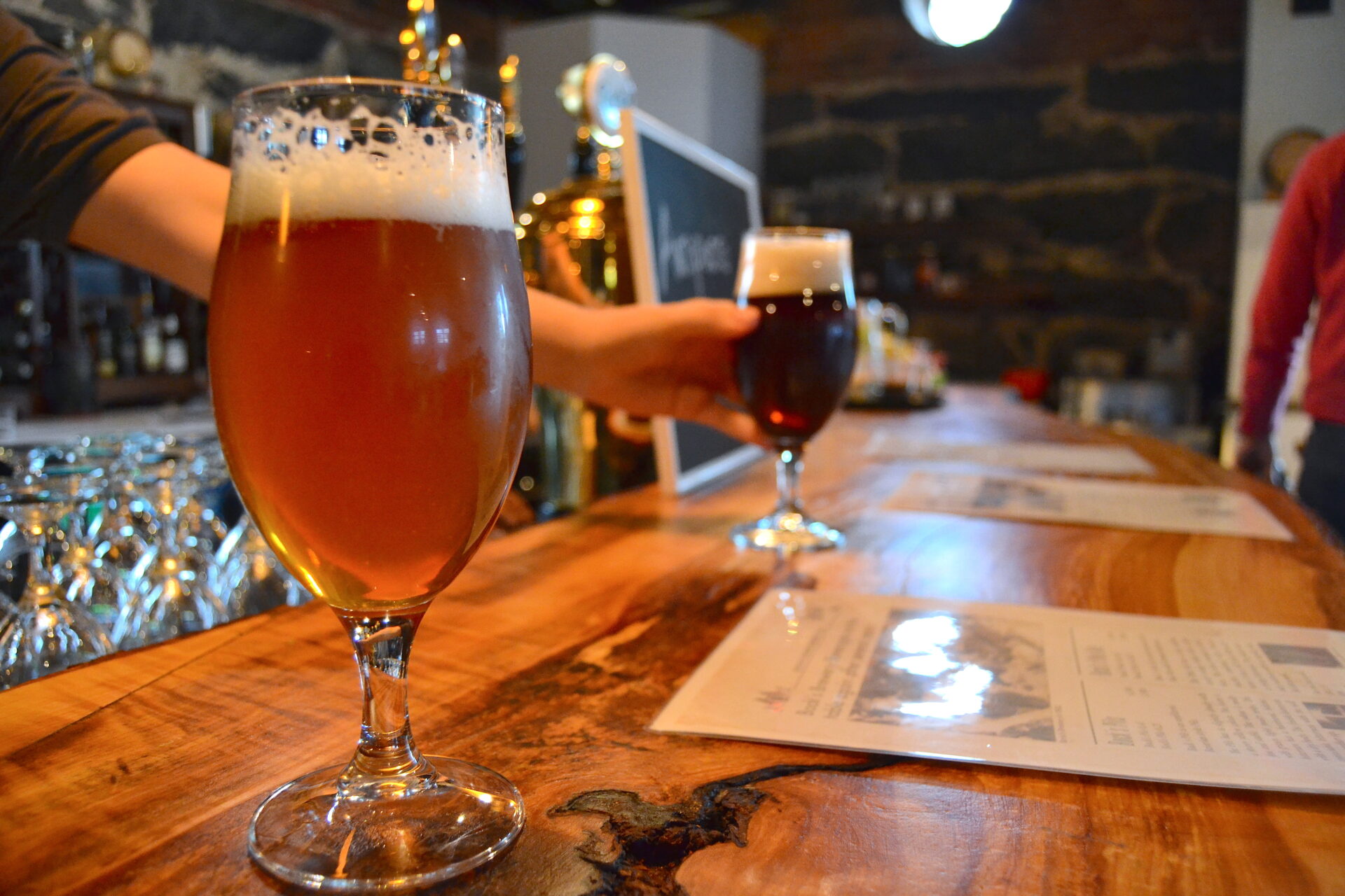 Close-up of a bar counter with two beer glasses. In the foreground is a light beer, and in the background, a dark beer is being handed over.