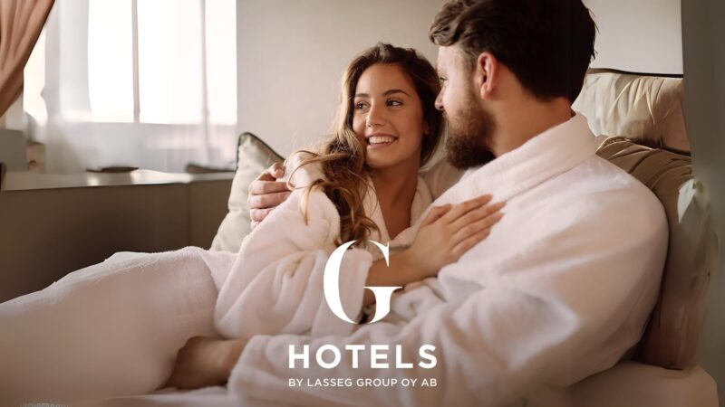 Kaksi henkilöä kylpytakeissa istuvat sohvalla ja hymyilevät toisilleen. Kuvassa on G Hotels -logo.