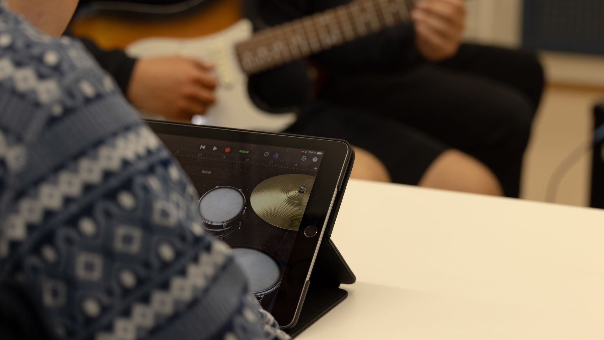 A person is looking at a tablet displaying drums on the screen. In the background, another person is playing guitar.