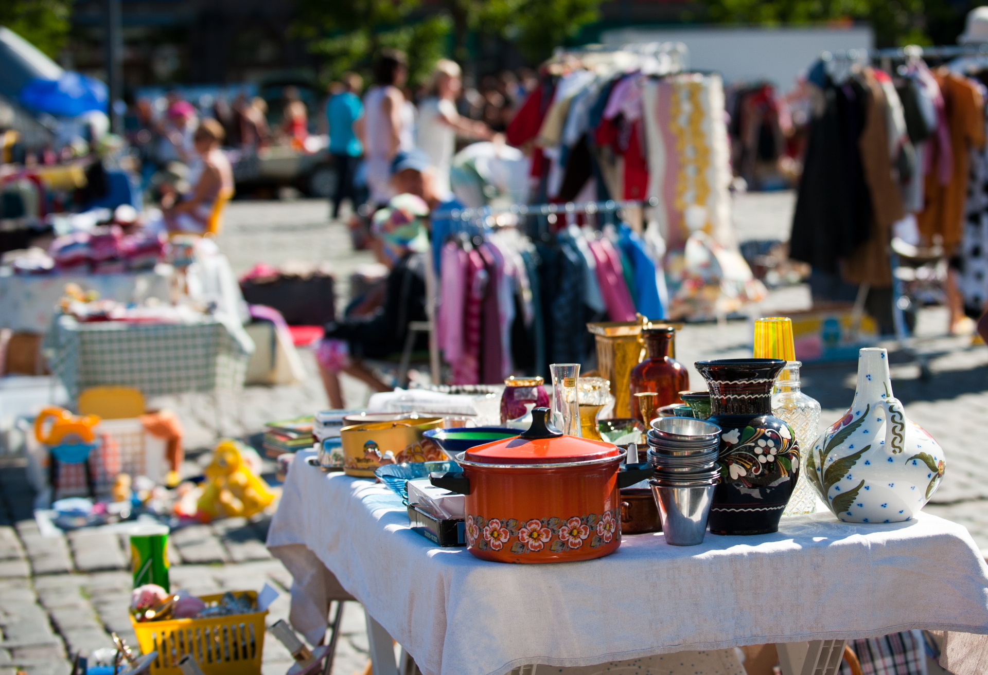 A flea market with tables full of various tableware and objects, with clothes hanging in the background.