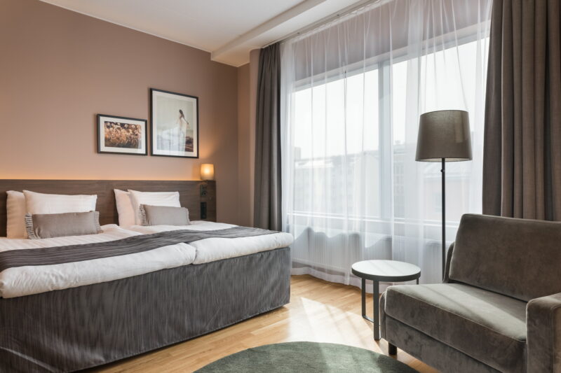 Hotellihuone, jossa on leveä sänky, kaksi taulua seinällä, nojatuoli, pyöreä pöytä ja suuri ikkuna.
