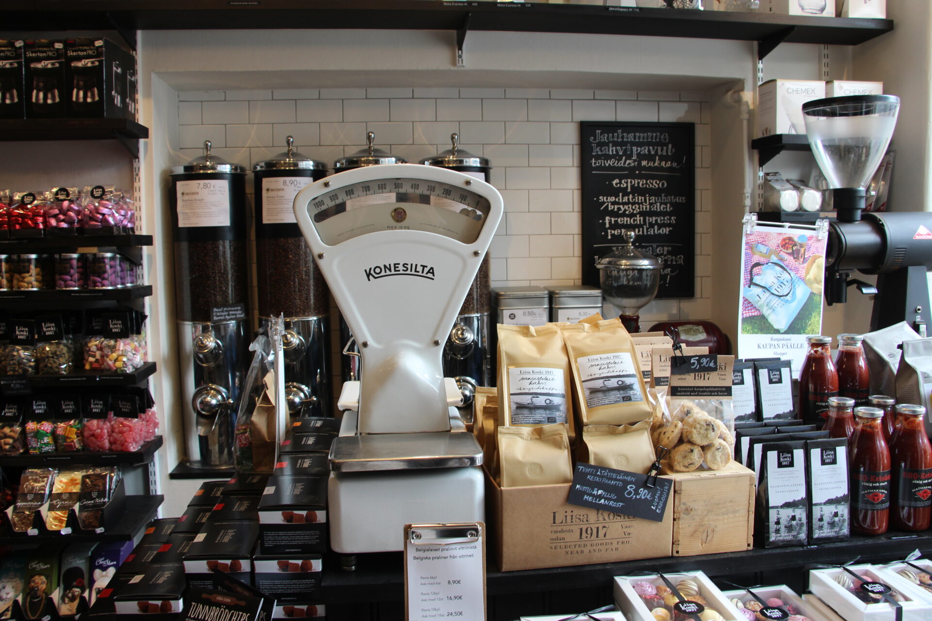 A coffee shop counter with a retro-style scale, bags of coffee beans and various treats. White tiles on the wall and a list of the coffees available.