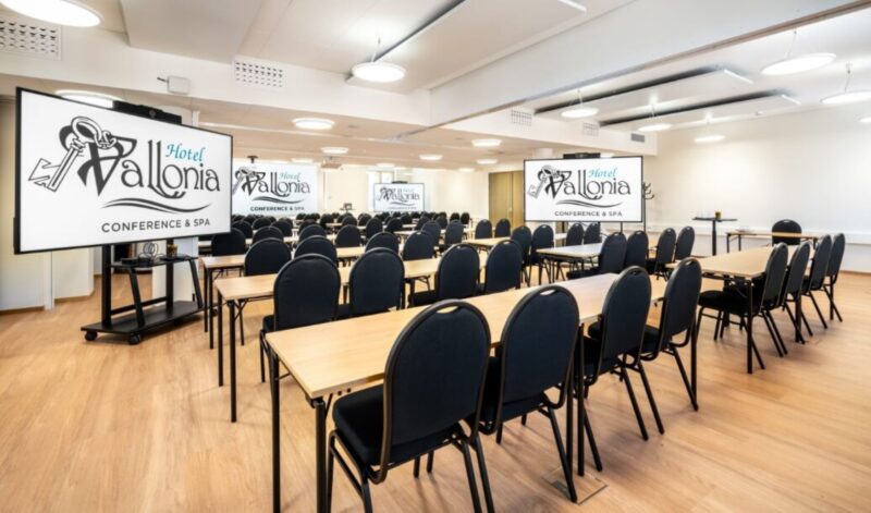 Conference room with several rows of tables and chairs, and a large screen displaying 'Hotel Vallonia Conference & Spa'.