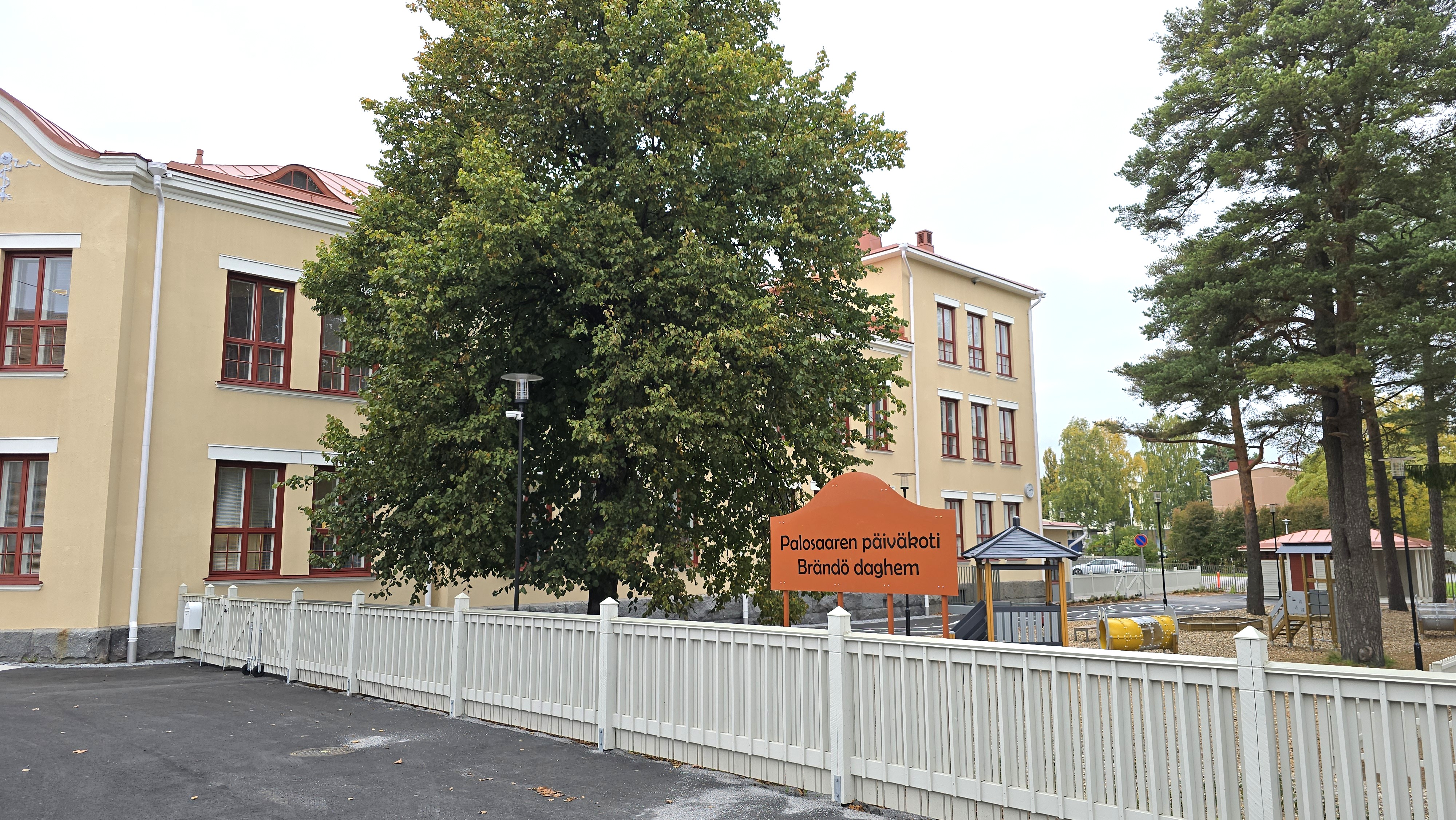 Bilden visar en ljus tvåvåningsbyggnad med ett vitt staket och ett stort träd framför. I förgrunden finns en skylt med texten 'Brändö daghem'.