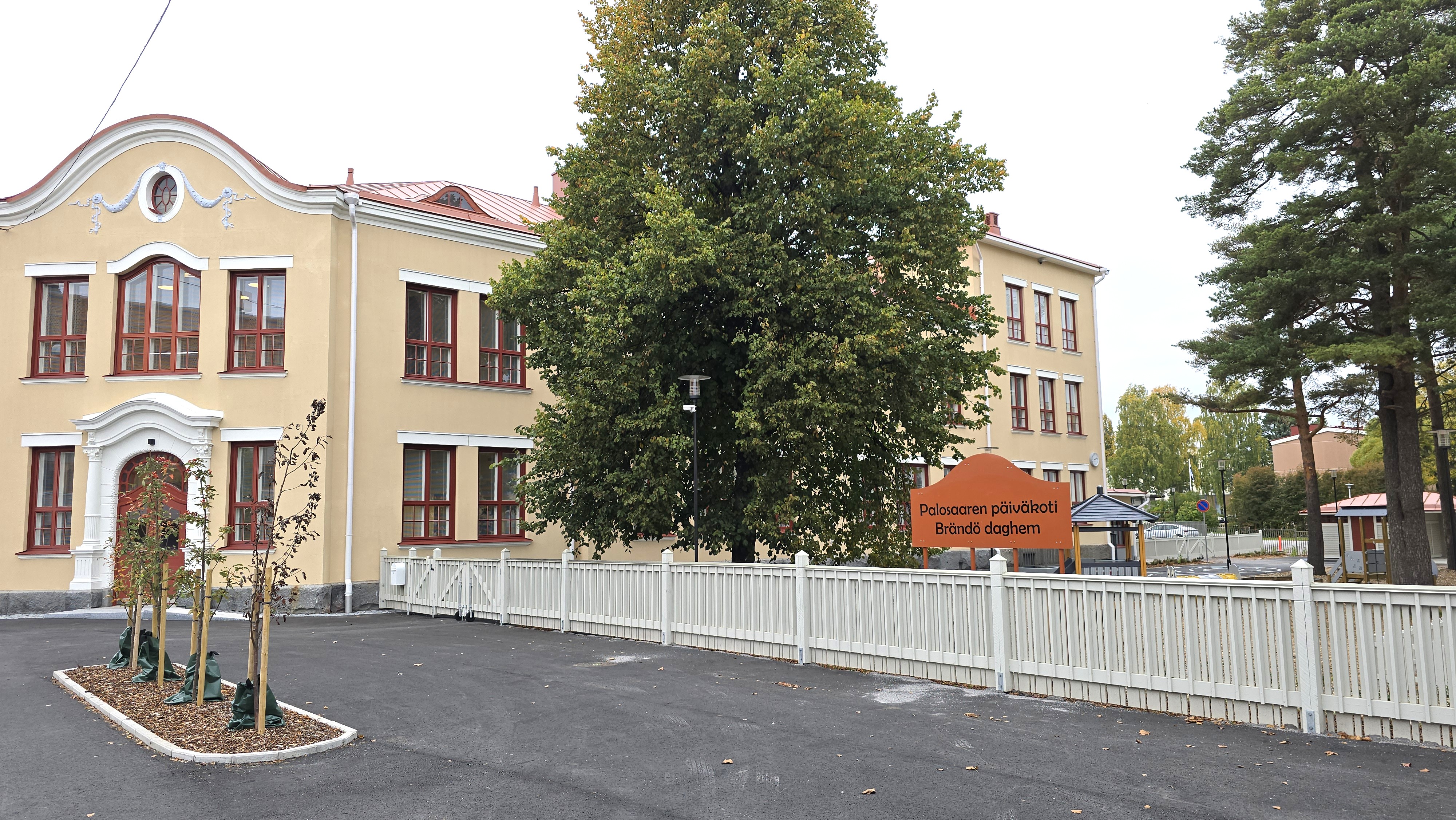 En byggnad med gammaldags utseende, gula väggar och röda fönsterkarmar. I förgrunden finns ett vitt staket och en skylt där det står 'Brändö daghem'.