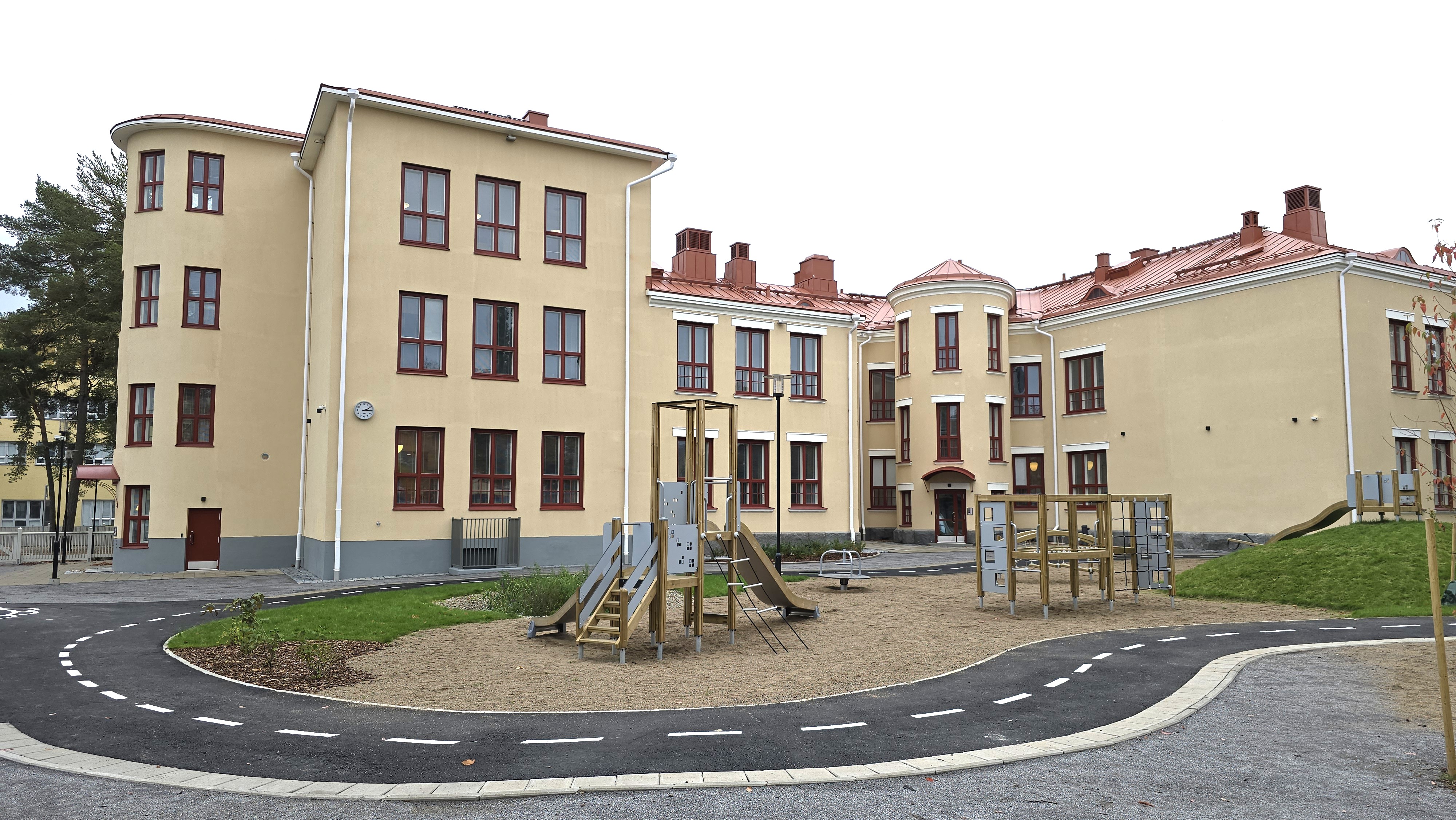 Bild på ett ljusgult daghem med en lekplats framför, med klätterställningar och sandfält.