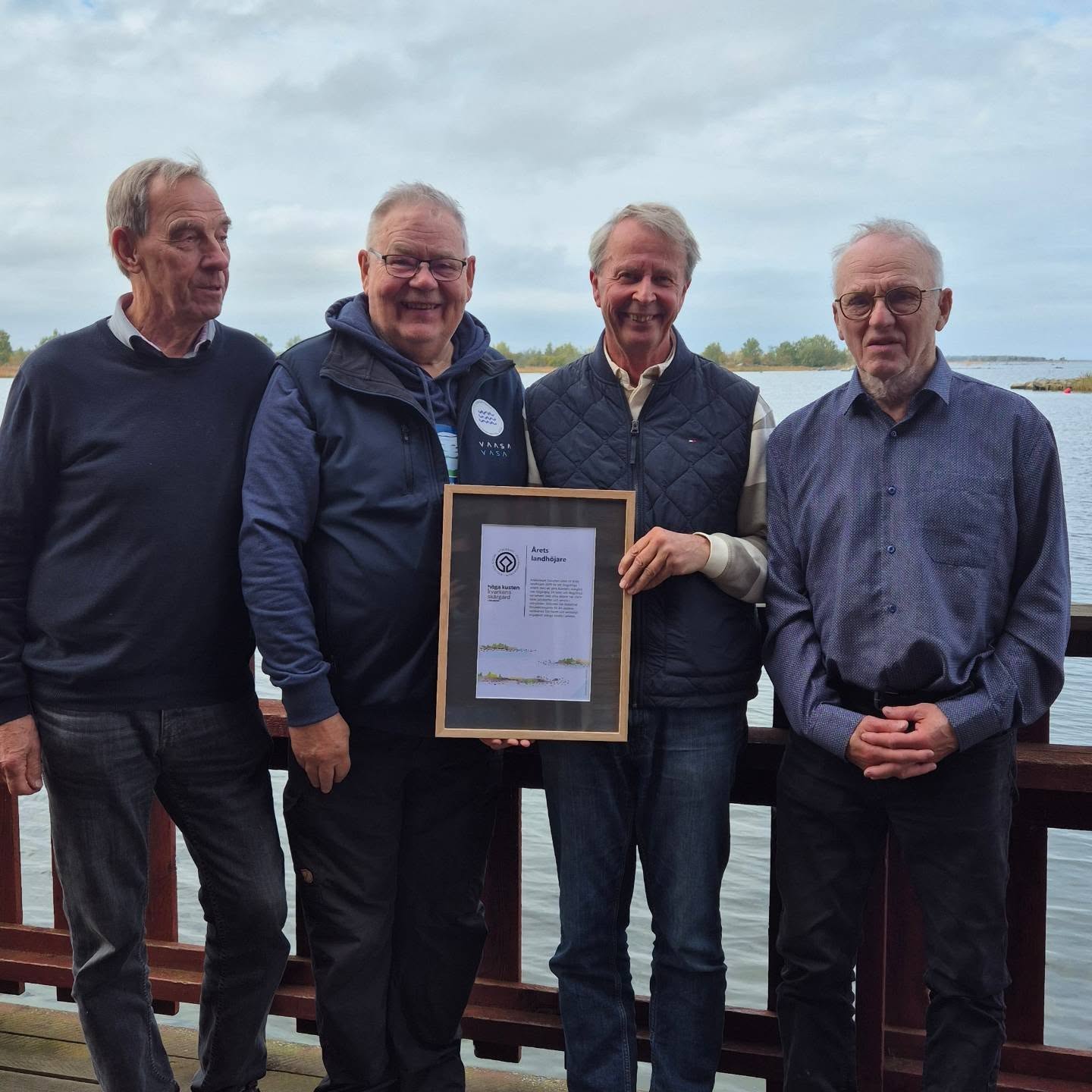 Four men stand outside on a jetty by the water, one of them holding a framed award.