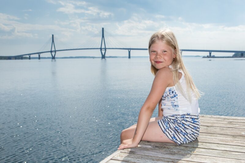 En ung flicka sitter på en brygga vid vattnet med en bro i bakgrunden.