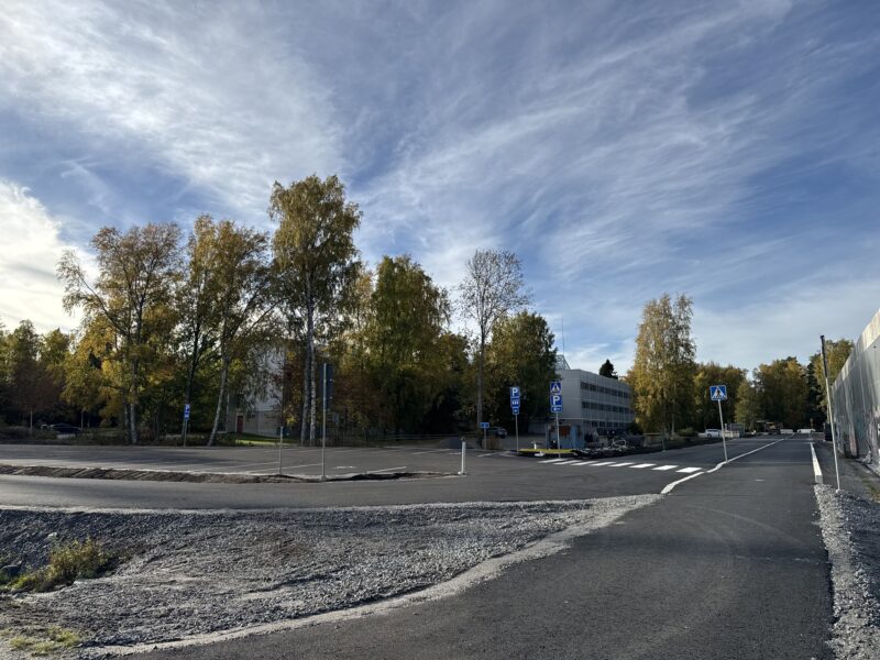 Syksyinen maisema, jossa on puiden reunustama tie ja rakennus taustalla. Taivas on pilvinen ja kirkas.