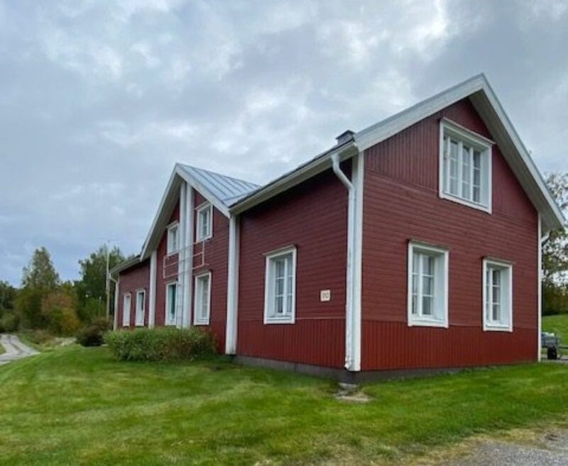 Punainen puutalo nurmikon ja tien vieressä pilvisen taivaan alla.