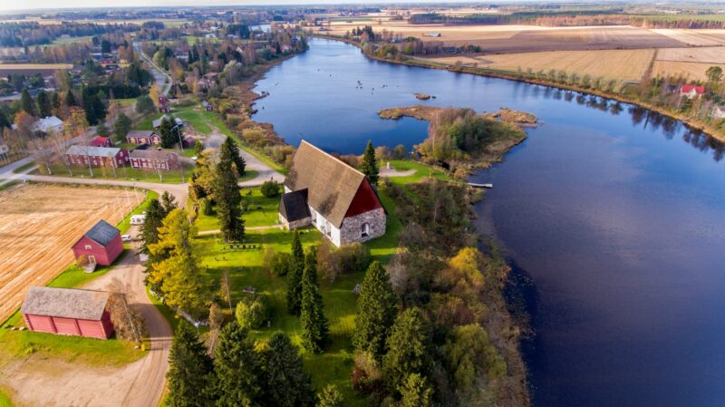 Flygfoto av ett landskap med en gammal stenkyrka och en flod. Runt kyrkan finns gröna träd och ängar.