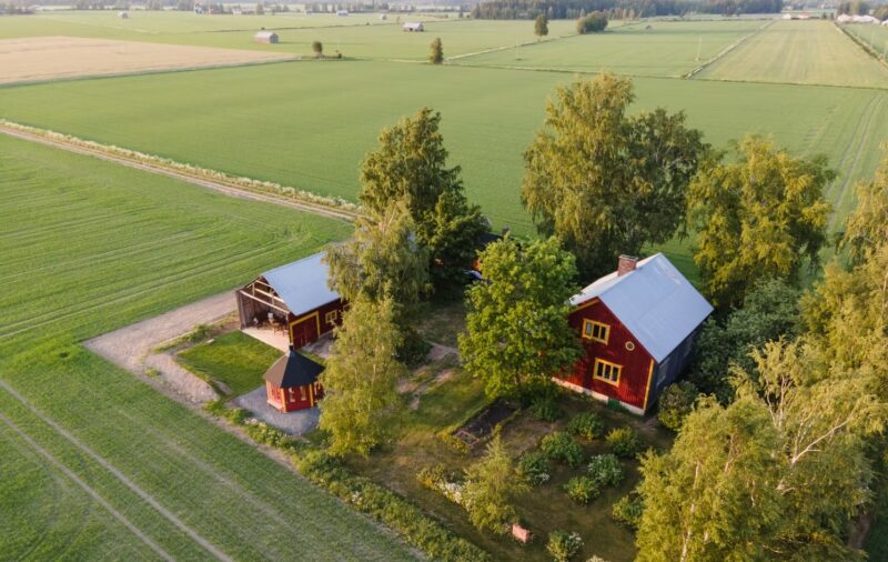 Ilmakuva maaseudun talosta ja navetasta, joita ympäröivät pellot ja puut.