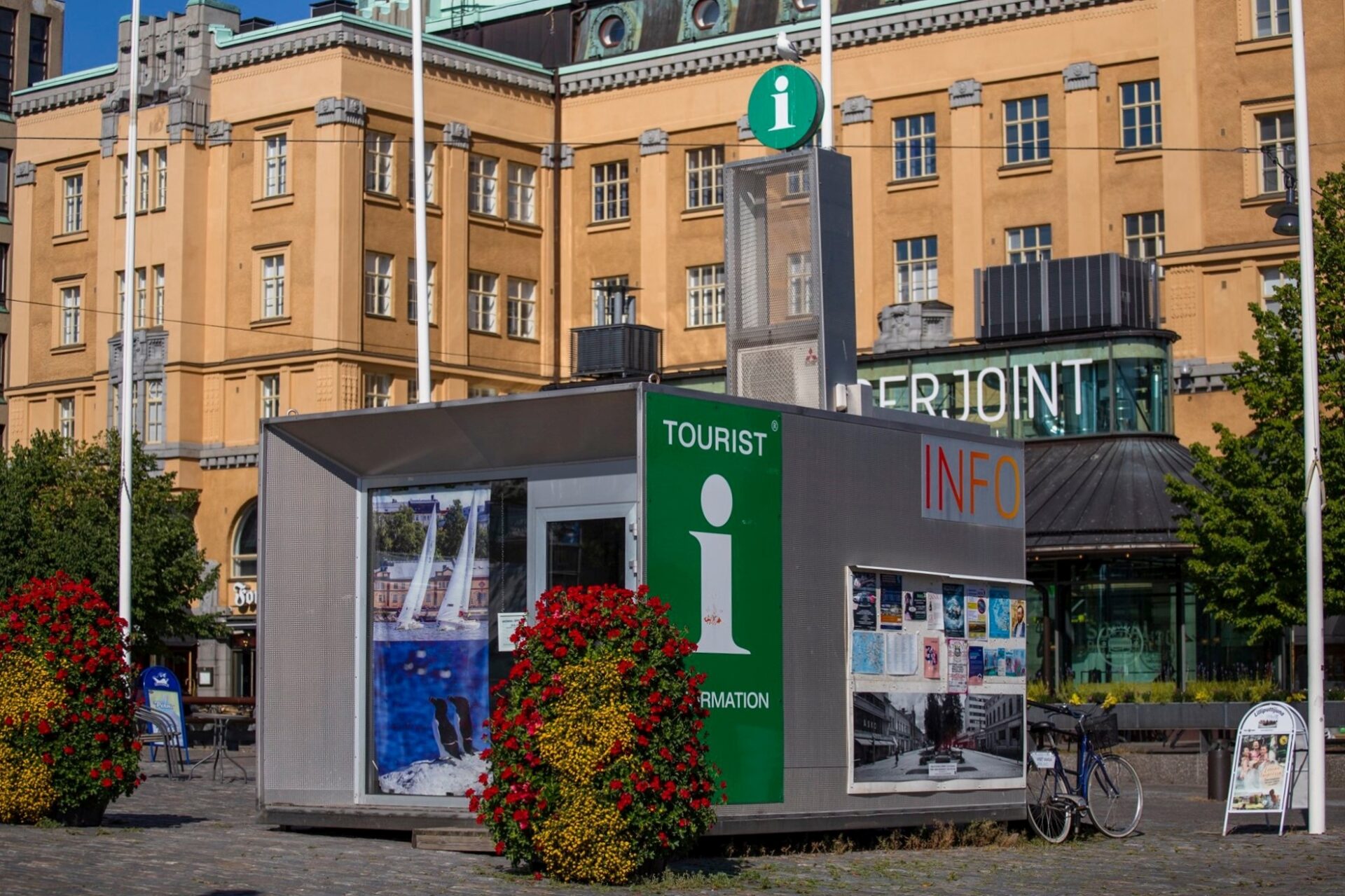 Image of a tourist information center in a city. In front of the building are round flower arrangements and in the background, there is a large old building.