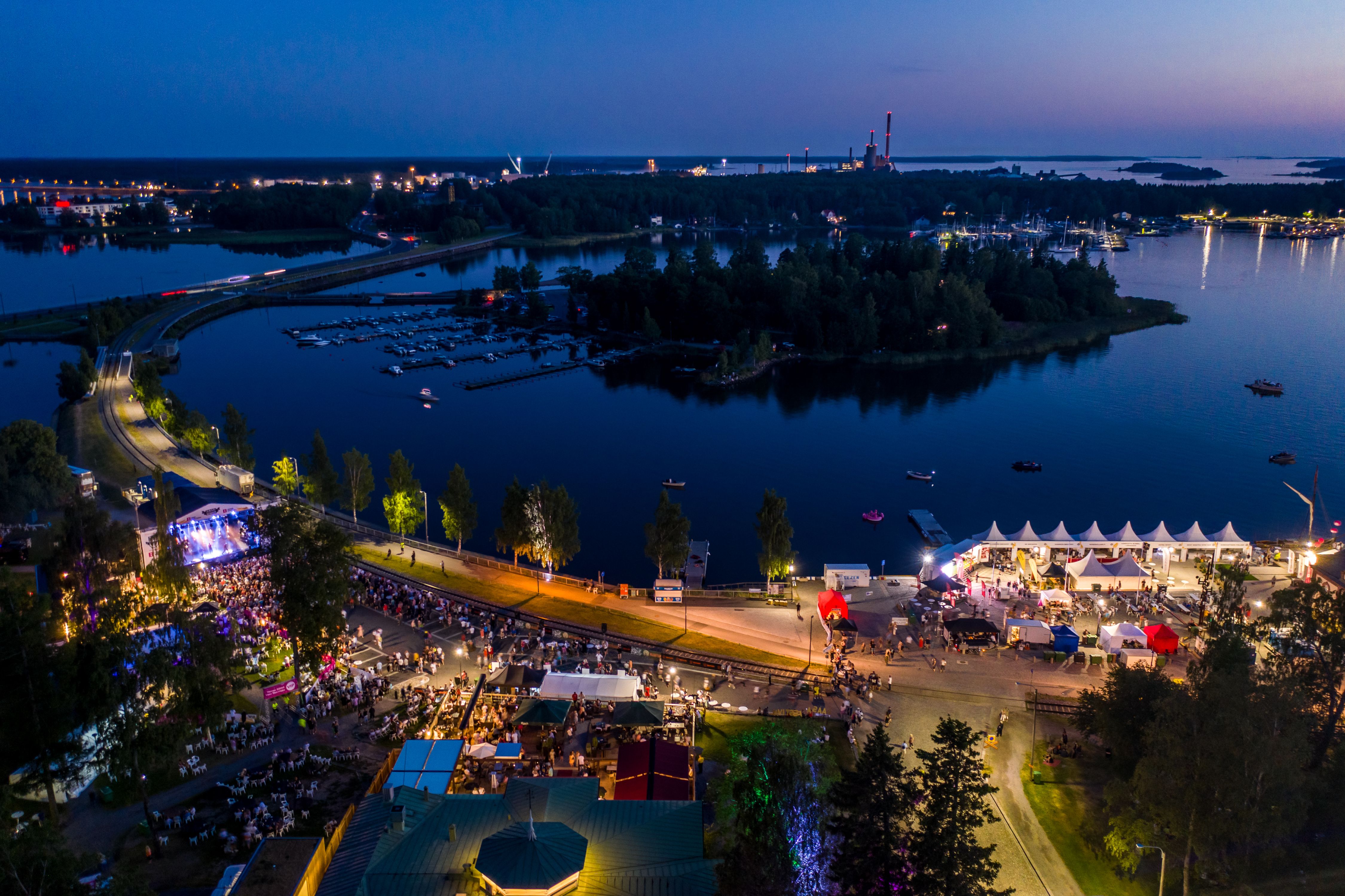 Aerial view of a festival at night by the water. Crowds, tents, and a lit-up stage are visible.
