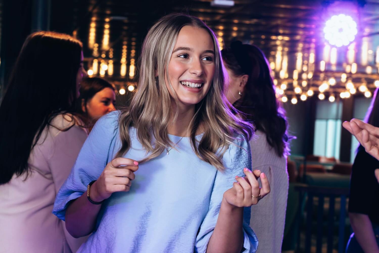 A young woman smiling and dancing at a party in a blue dress, with colorful lighting in the background.